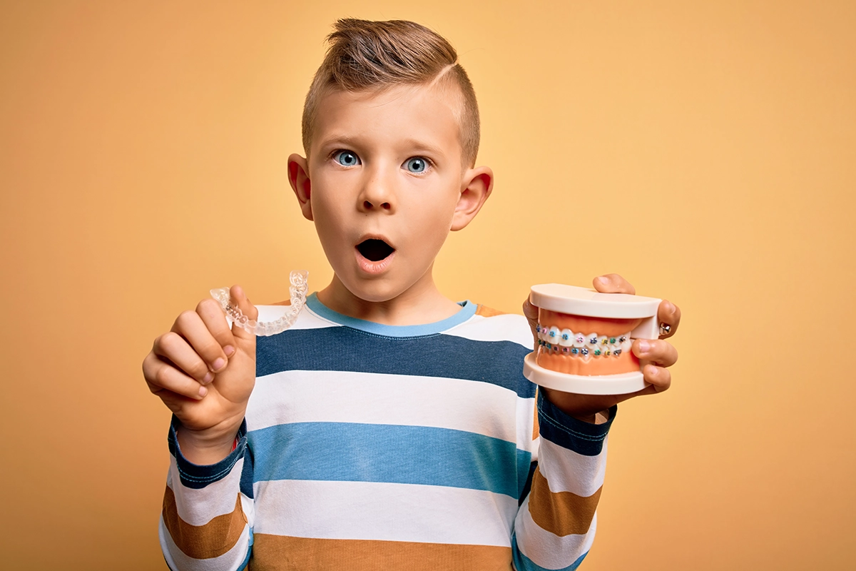 A surprised child holds a clear dental retainer in one hand and a model of teeth with braces in the other, against a warm orange background.